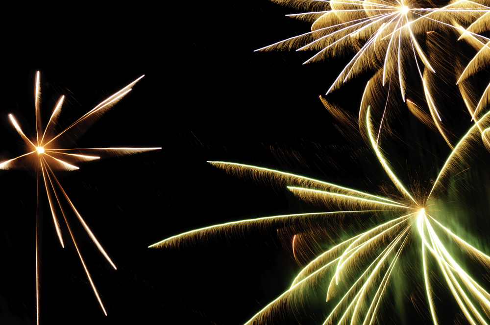 Bursts of fireworks with white-hot cores and reddish-orange, greenish, and white streaks with feathery motion blur